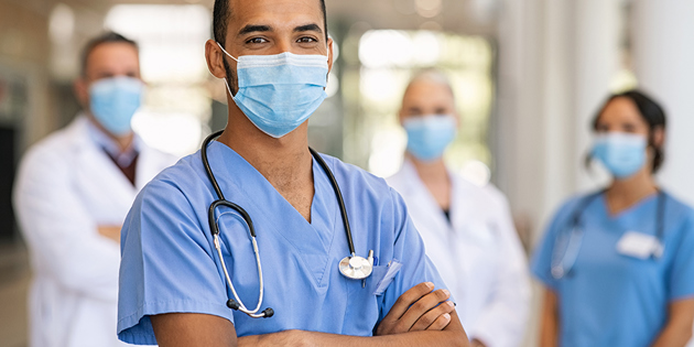Confident multiethnic male nurse in front of his medical team looking at camera wearing face mask during covid-19 outbreak. Happy and proud indian young surgeon standing in front of his colleagues wearing surgical mask for prevention against coronavirus. Portrait of mixed race doctor with medical staff in background at hospital.